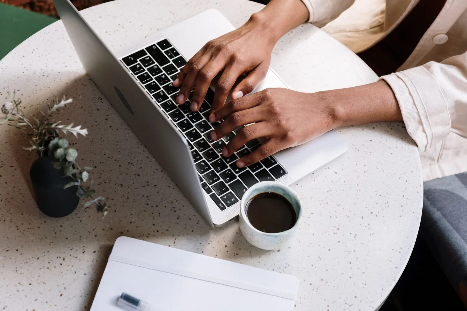 Hands typing on a laptop at a cafe table with a notebook and coffee, writing long-form content